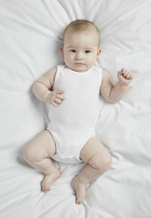 Weißer Baumwoll-Overall auf einem Baby, das auf einer weißen Stoffoberfläche liegt. Das Baby hat helle Haut, kurzes braunes Haar und zeigt einen neutralen Gesichtsausdruck.