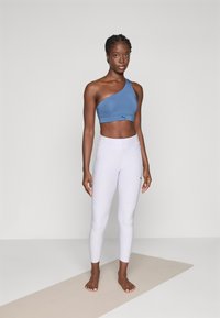 Vrouw staat blootsvoets op een beige yogamat, gekleed in een blauwe sportbeha met één schouder en witte high-waisted legging, tegen een neutrale achtergrond.