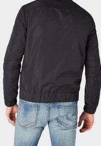 Giacca bomber leggera nera da uomo con polsini elasticizzati, caratterizzata da un'esterno liscio e una finitura leggermente strutturata; indossata sopra jeans azzurri chiari.