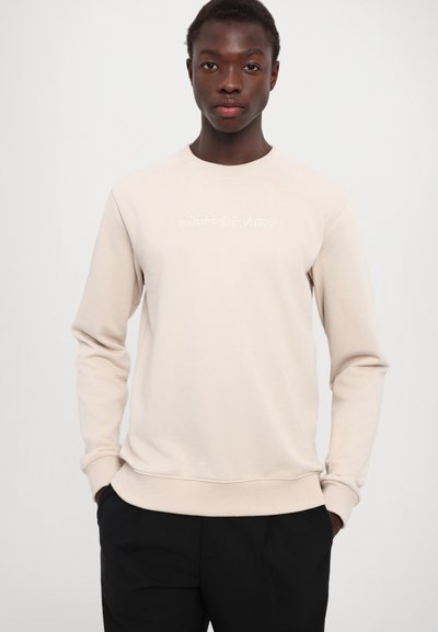 Lys beige genser med lange ermer, ribbestrikkede mansjetter og kant. Har hvit tekst "#HOUSE OF JAFFA" på brystet. Glatt stofftekstur.