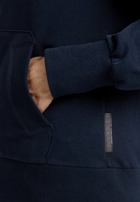 Námořnická modrá mikina vyrobená z měkkého materiálu, která má klokaní kapsu, žebrované manžety a texturovanou nášivku na přední straně.