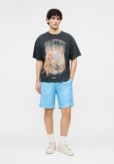 Jeune homme aux cheveux bouclés portant un t-shirt graphique noir, un short bleu clair, des chaussettes blanches et des baskets beiges, debout avec une main dans la poche.