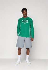 Zelena majica z dolgimi rokavi z napisom "Celtics" in logotipom Nike, kombinirana s svetlo sivimi kratkimi hlačami in belimi supergami. Preprost športni dizajn.
