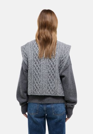 Personne aux longs cheveux châtain clair portant un gilet en maille torsadée gris par-dessus un pull gris et un jean bleu, dos tourné sur un fond uni.