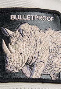 Baseball cap featuring a black patch with an embroidered rhinoceros and the word "BULLETPROOF." Cap is beige with a structured shape.