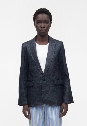 Personne portant un blazer en cuir noir froissé sur une chemise blanche et un pantalon à rayures verticales bleu, jaune et blanc sur un fond uni.