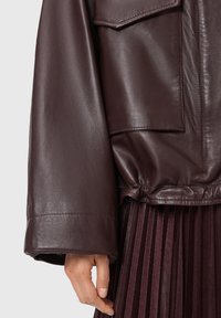Mano appoggiata di lato, indossando una giacca in pelle marrone scuro con maniche larghe e una gonna plissettata color borgogna sotto.