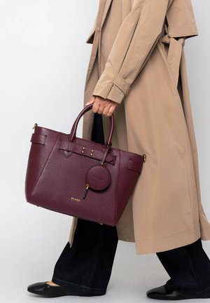 Burgundy leather handbag with textured surface, dual handles, gold-tone hardware, and a detachable round tag. Worn under a beige coat.