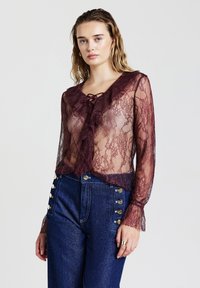 Blusa in pizzo color bordeaux con scollo a V, davanti arricciato e fiocco. Presenta tessuto trasparente e maniche lunghe. Abbinata a denim a vita alta.