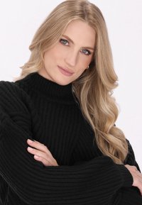 Czarny sweter prążkowany z wysokim kołnierzem, charakteryzujący się miękką teksturą i luźnymi rękawami. Modelka ma długie, falujące blond włosy.