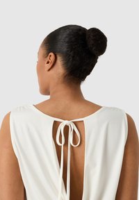 Femme aux cheveux bruns attachés en chignon, portant un haut sans manches blanc avec un dos ouvert noué par un nœud à la nuque.