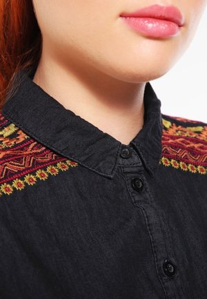 Primo piano della parte inferiore del volto e del collo di una persona che indossa una camicia nera con bottoni e motivi ricamati rossi e gialli sulle spalle.