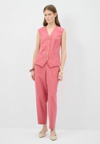 Blazer sans manches rose avec des boutons dorés et deux poches, associé à un pantalon taillé assorti, complété par des talons hauts de couleur claire.
