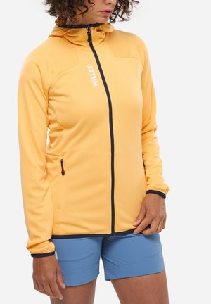 Femme portant une veste zippée jaune à capuche et un short bleu, debout devant un fond blanc uni.