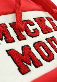 Hoodie rouge et blanc avec texte brodé "Mickey Mouse", présentant des lettres texturées et des contours noirs contrastants. Cordon de serrage ajustable pour la capuche.