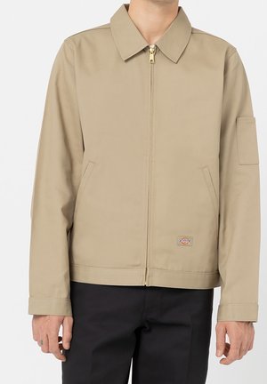 Person trägt eine beige Jacke mit Reißverschluss und Kragen, zwei Fronttaschen und einem kleinen Logo-Aufnäher nahe der Taille, kombiniert mit einer schwarzen Hose.