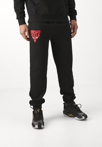 Pantalones de chándal negros con un gráfico rojo de "Chicago Bulls" en el muslo izquierdo, con un corte relajado y tobillos elásticos. Combinados con zapatillas negras.