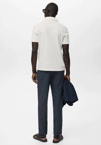 White polo shirt, dark trousers, and sandals. Individual holds a navy jacket. Clean lines, simple design, modern materials.