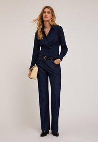 Camicia a maniche lunghe blu navy, pantaloni in denim scuri a vita alta, cintura con catena, borsa color crema, abbinati a décolleté nere con punta appuntita.