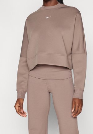 Kobieta nosząca dopasowaną taupe krótką bluzę i legginsy z wysokim stanem z małym białym logo Nike na piersi bluzy.