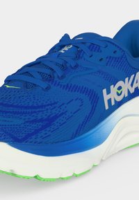 Μπλε παπούτσι για τρέξιμο Hoka με υφασμάτινο άνω μέρος με υφή, λευκή σόλα με μπλε διαβάθμιση, εξάρσεις σε πράσινο λάιμ στην εξωτερική σόλα και λευκό λογότυπο Hoka στο πλάι.