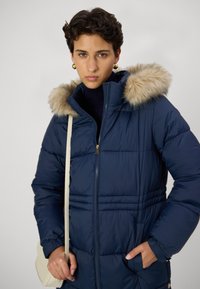 Jūras spēku puffer jaka ar mākslīgās ādas kapuci, horizontālu šuvju, zelta rāvējslēdzēju un sānu kabatām, apvienota ar vieglu plecu somu.