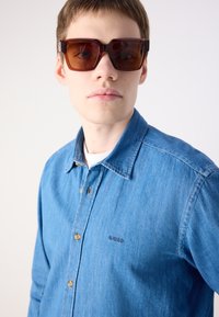 Camicia in denim azzurro chiaro con colletto a punta e bottoni marroni, caratterizzata da una sottile ricamo "BOSS" sul lato sinistro del petto e maniche lunghe.