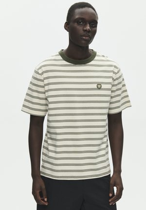 Ung mand iført en cremefarvet T-shirt med korte ærmer med tynde sorte, vandrette striber og et lille olivengrønt logo på venstre bryst.