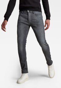 Jeans de mezclilla gris con un corte slim, diseño de cinco bolsillos y cremallera. Combinados con zapatillas blancas, mostrando un look casual.