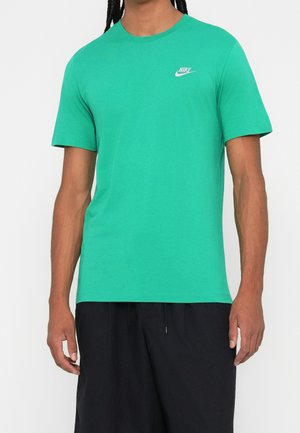 Homme portant un t-shirt Nike vert vif à manches courtes et un short noir, debout devant un fond blanc uni.