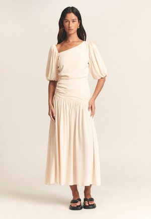 Lichtbeige, een-schouder jurk met pofmouwen, geraffineerde taille en vloeiende rok, gemaakt van zachte stof. Gecombineerd met zwarte platform sandalen.