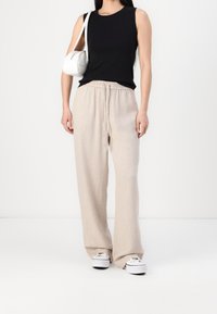 Mujer con una blusa sin mangas negra, pantalones beige de pierna ancha, zapatillas blancas y un bolso blanco en su brazo derecho.