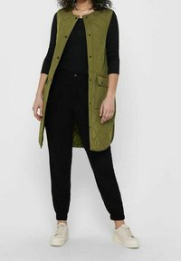 Pikowana oliwkowo-zielona kamizelka z zatrzaskami, bocznymi kieszeniami i długim fasonem, w zestawieniu z czarnymi spodniami i białymi trampkami.