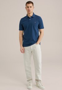 Mornarska polo majica s kratkimi rokavi, ovratnikom in logotipom na levi strani prsnega dela, v kombinaciji s svetlimi, ohlapnimi hlačami in belimi supergami.