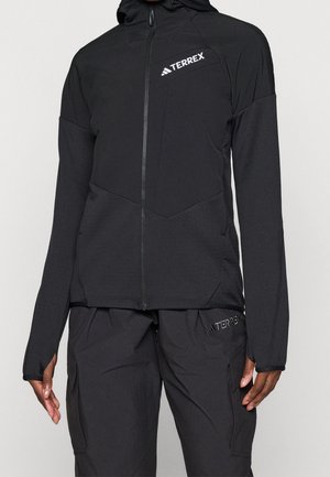 Personne portant une veste à capuche noire zippée et un pantalon noir avec des logos Terrex sur la poitrine et la cuisse.