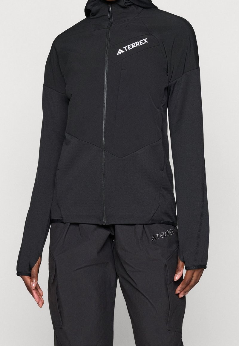 Personne portant une veste à capuche noire zippée et un pantalon noir avec des logos Terrex sur la poitrine et la cuisse.