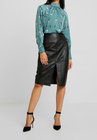 Blusa di stampa floreale color teal con scollatura alta e maniche a palloncino, abbinata a una gonna a tubino in pelle nera con spacco frontale. Tacchi neri.