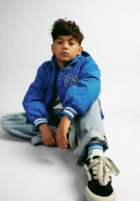 Giubbotto bomber blu con polsini a strisce bianche, indossato sopra una camicia bianca, abbinato a jeans blu chiaro e sneakers in bianco e nero.