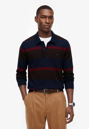 Mann trägt ein navyblau, schwarz und rot gestreiftes langärmeliges Poloshirt, beige Hose und eine silberne Armbanduhr, steht mit einer Hand in der Tasche.