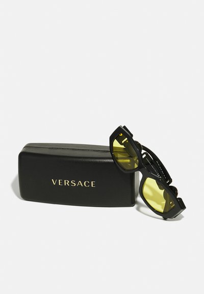 Óculos de sol Versace com armação preta, lentes amarelas e logótipo dourado. Acompanhados por uma caixa retangular preta.