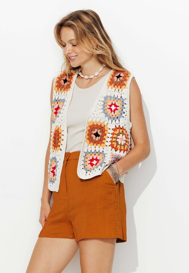 Gilet au crochet mettant en avant des carrés granny multicolores. Base blanche avec des accents orange, bleu et rose. Associé à un short rouille. Design décontracté.