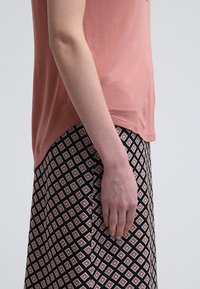 Vue de profil d'une personne portant un haut rose à manches courtes et une jupe noire avec des motifs losanges roses et blancs, bras détendu le long du corps.