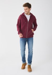 Veste à capuche bordeaux zippée avec cordon de serrage, deux poches avant, portée sur un t-shirt blanc, associée à un jean bleu clair et des bottes beiges.