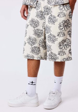 Persona che indossa pantaloncini bianchi fino al ginocchio con motivi floreali astratti neri, calzini bianchi con logo nero e sneakers bianche, in piedi contro uno sfondo semplice.