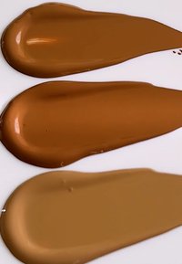 Tre fargeprøver av flytende foundation i forskjellige nyanser av varm brun, med glatte, blanke teksturer på hvit bakgrunn.