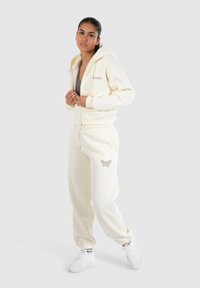 Crèmekleurige rits hoodie met voorzakken en logo; bijpassende crème sweatpants met een decoratief vlinderontwerp; witte sneakers.