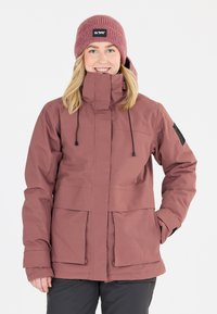 Wasserdichte pinke Jacke mit hohem Kragen, Reißverschluss vorne, verstellbaren Kordeln und zwei großen Taschen. Getragen mit einer passenden Strickmütze.