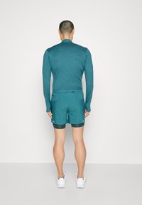 Teal långärmad tröja som matchas med teal shorts. Tröjan har en slät textur; shortsen har en lagerdesign. Fötterna pryds av vita sportskor.