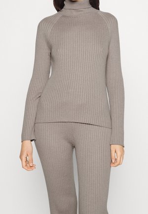 Femme portant un pull à col roulé en maille côtelée taupe et un pantalon assorti en maille côtelée, debout devant un fond uni.