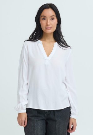 Mujer con cabello largo y negro, vistiendo una blusa blanca de manga larga con cuello en V y pantalones oscuros, de pie frente a un fondo liso.